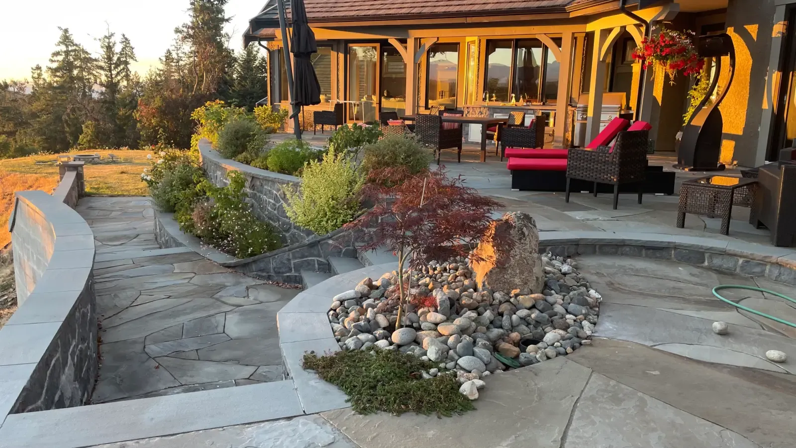 Completed multi-level flagstone patio with a curved stone planter, Japanese-maple feature bed, and furnished dining deck glowing at golden hour, overlooking the Strait of Georgia