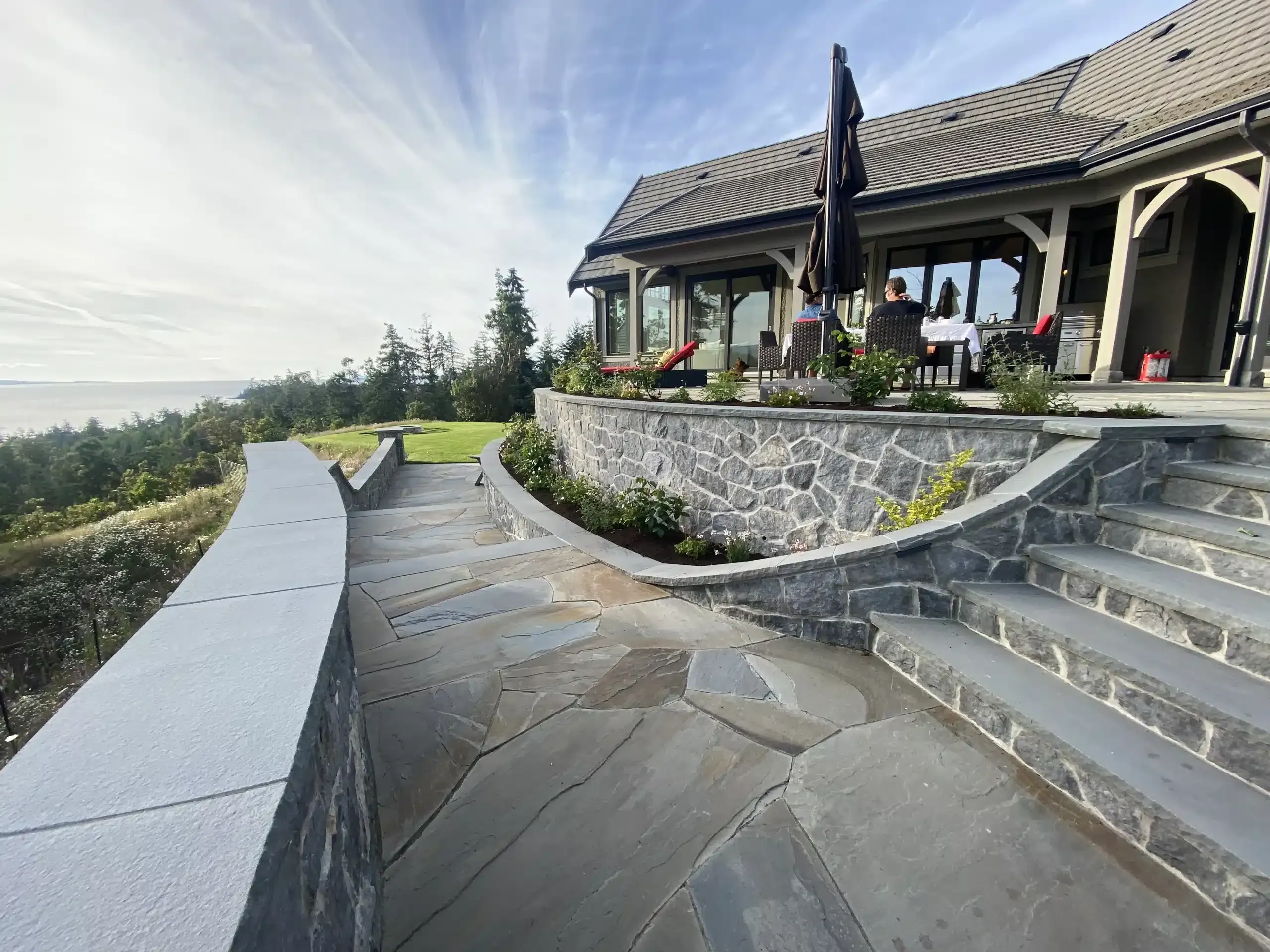 The completed flagstone patio and curved natural-stone retaining wall with fresh planting beds and a furnished covered deck