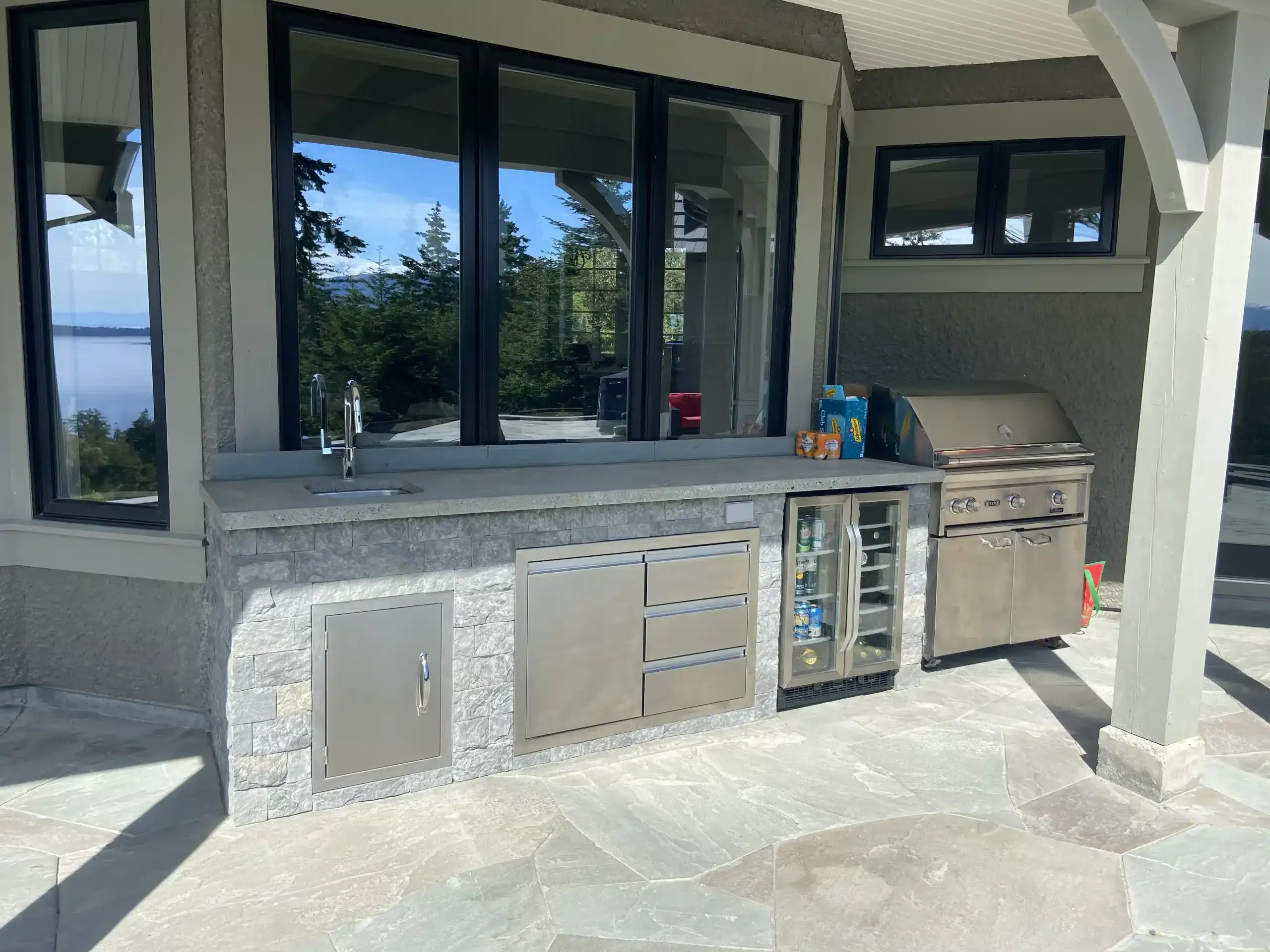 Finished covered outdoor kitchen with stone-faced cabinetry, built-in grill, beverage fridge, and ocean views framed in the windows