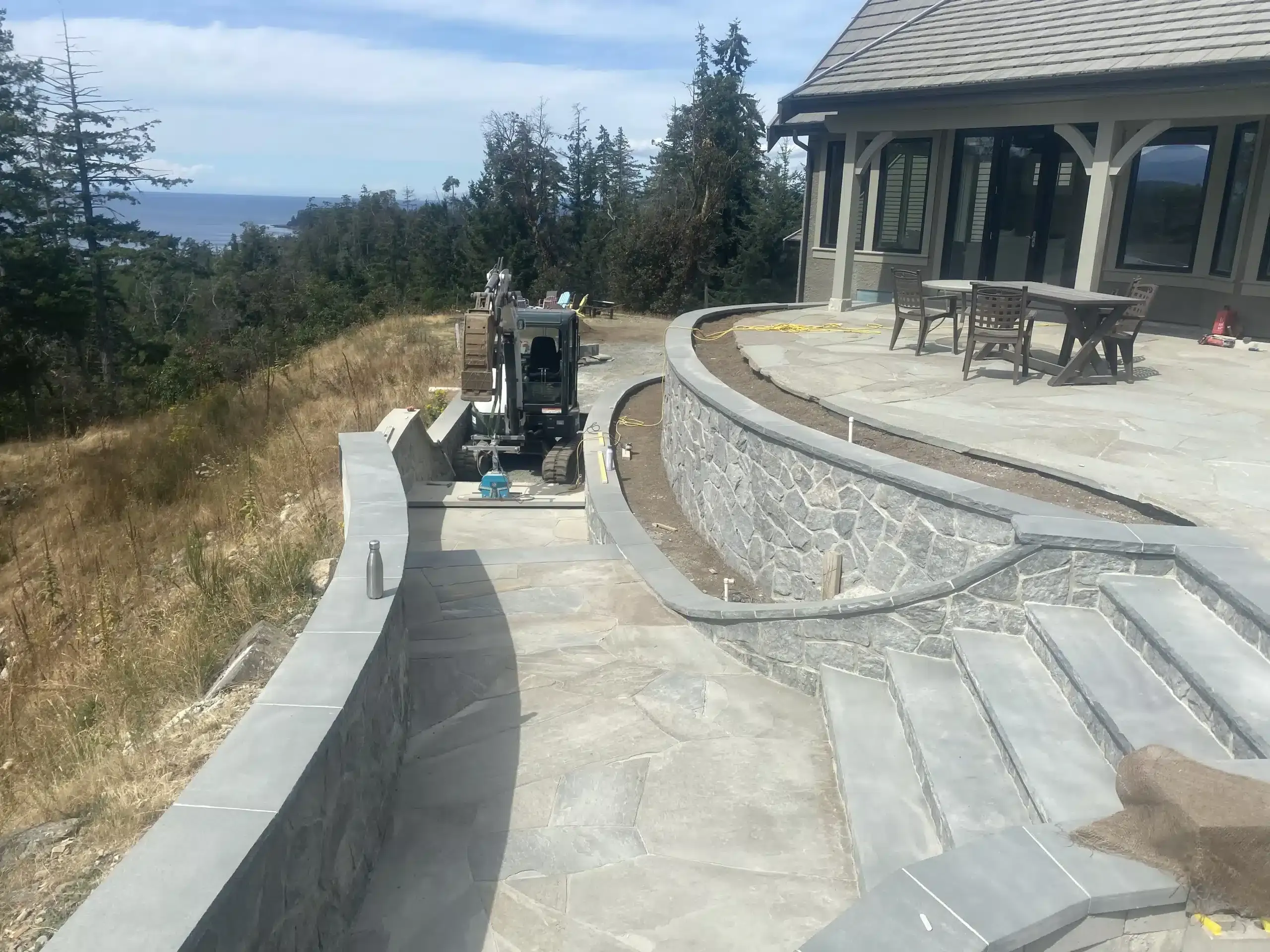 Curved gray stone retaining walls and flagstone patio under construction, with a mini-excavator working the wall